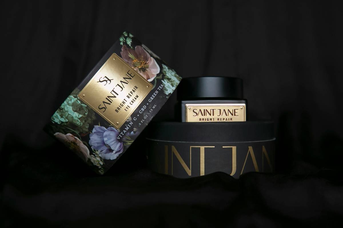 Beautiful floral on black with gold accent packing for an eye cream product called Saint Jane, Bright Repair Eye Cream, that is made from Vitamin C, Green Tea and CBD on a dark black background.